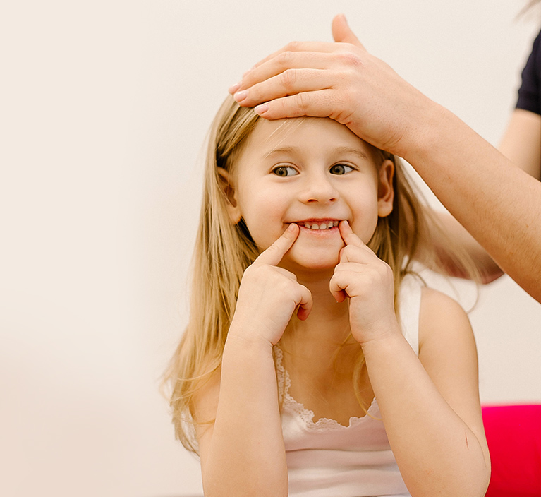 little girl smiling, holding two fingers to her bottom lip. An adult has their hand on her forehead.